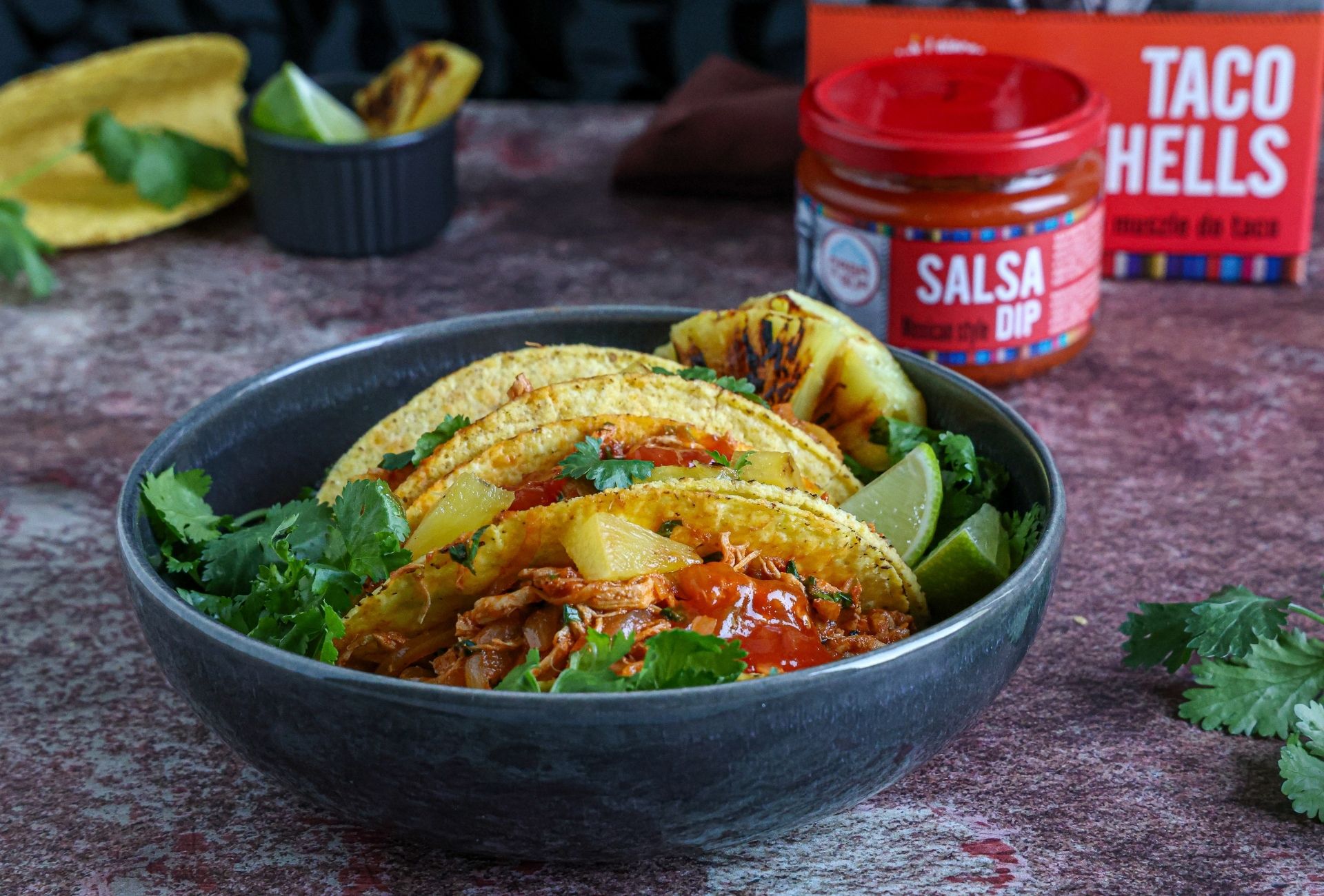 Tacos al Pastor z szarpaną wieprzowiną, ananasem i kolendrą w muszlach kukurydzianych w misce, podane obok słoiczka Salsa Dip oraz muszli Taco Shells marki Casa del Sur.