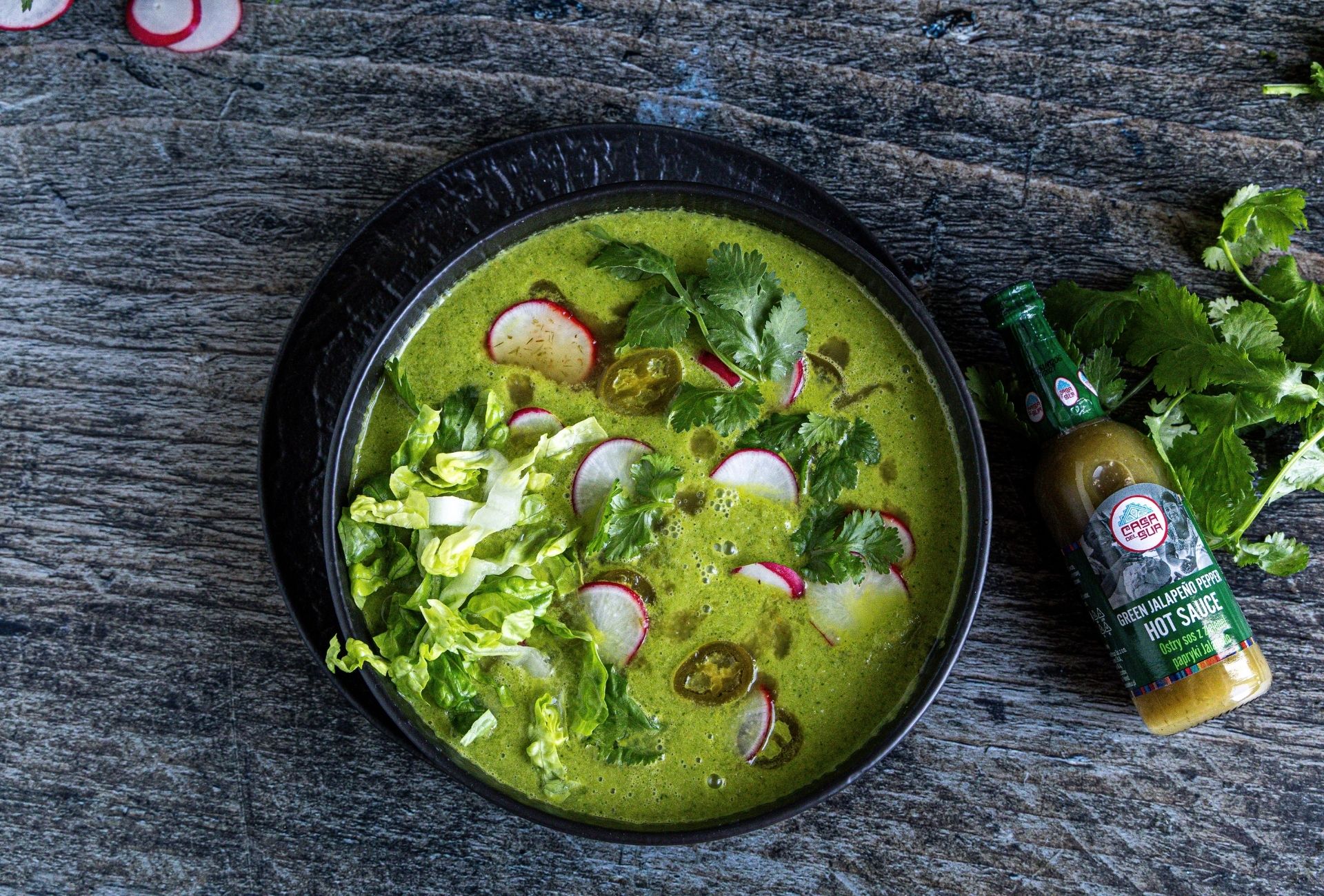 Pozole verde, sopa verde con rábano, lechuga y cilantro en un tazón negro, servida junto a una botella de salsa de jalapeño verde Casa del Sur.