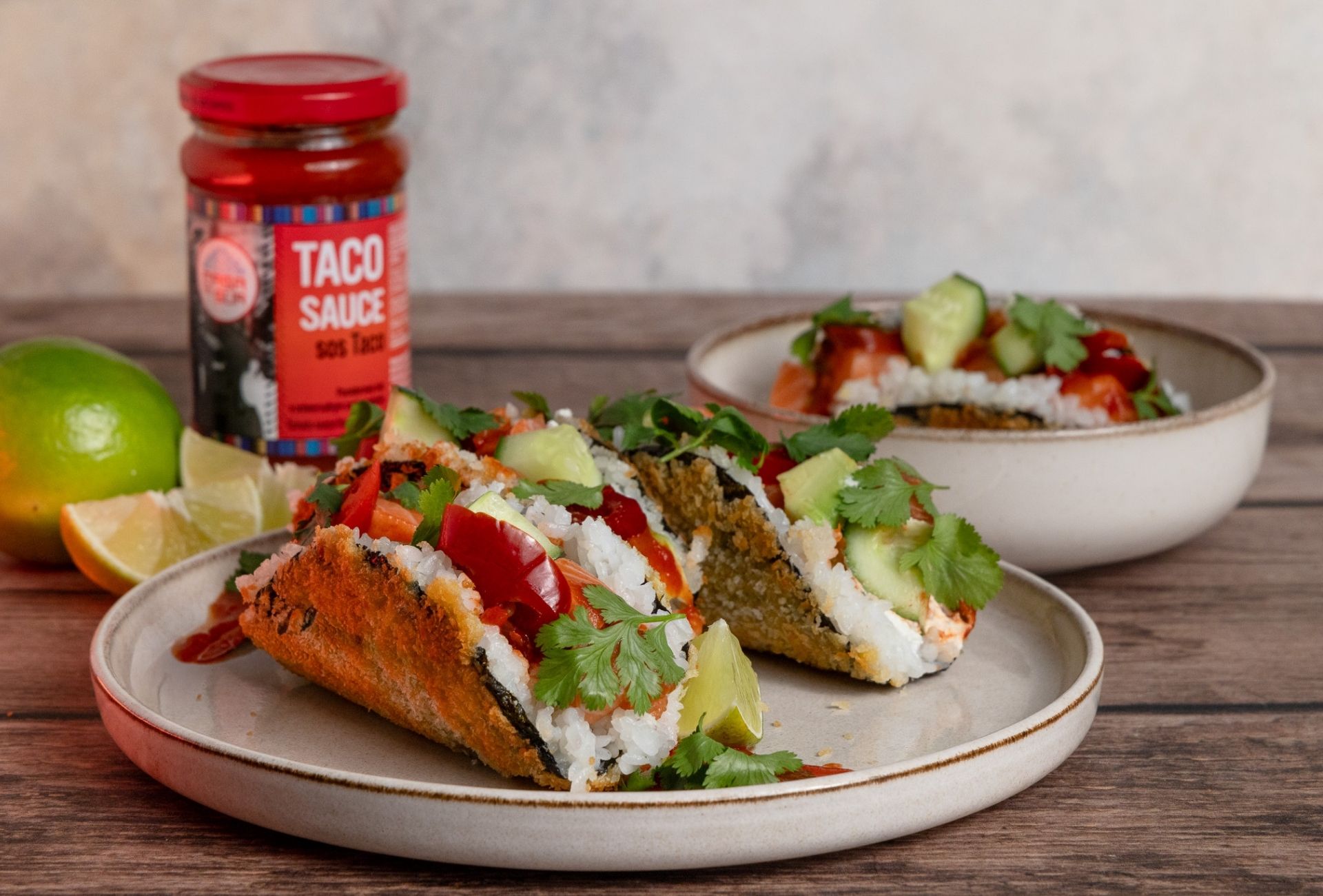 Taco-sushi z łososiem, ryżem, ogórkiem i kolendrą podane na talerzu obok słoiczka sosu taco marki Casa del Sur oraz limonki.