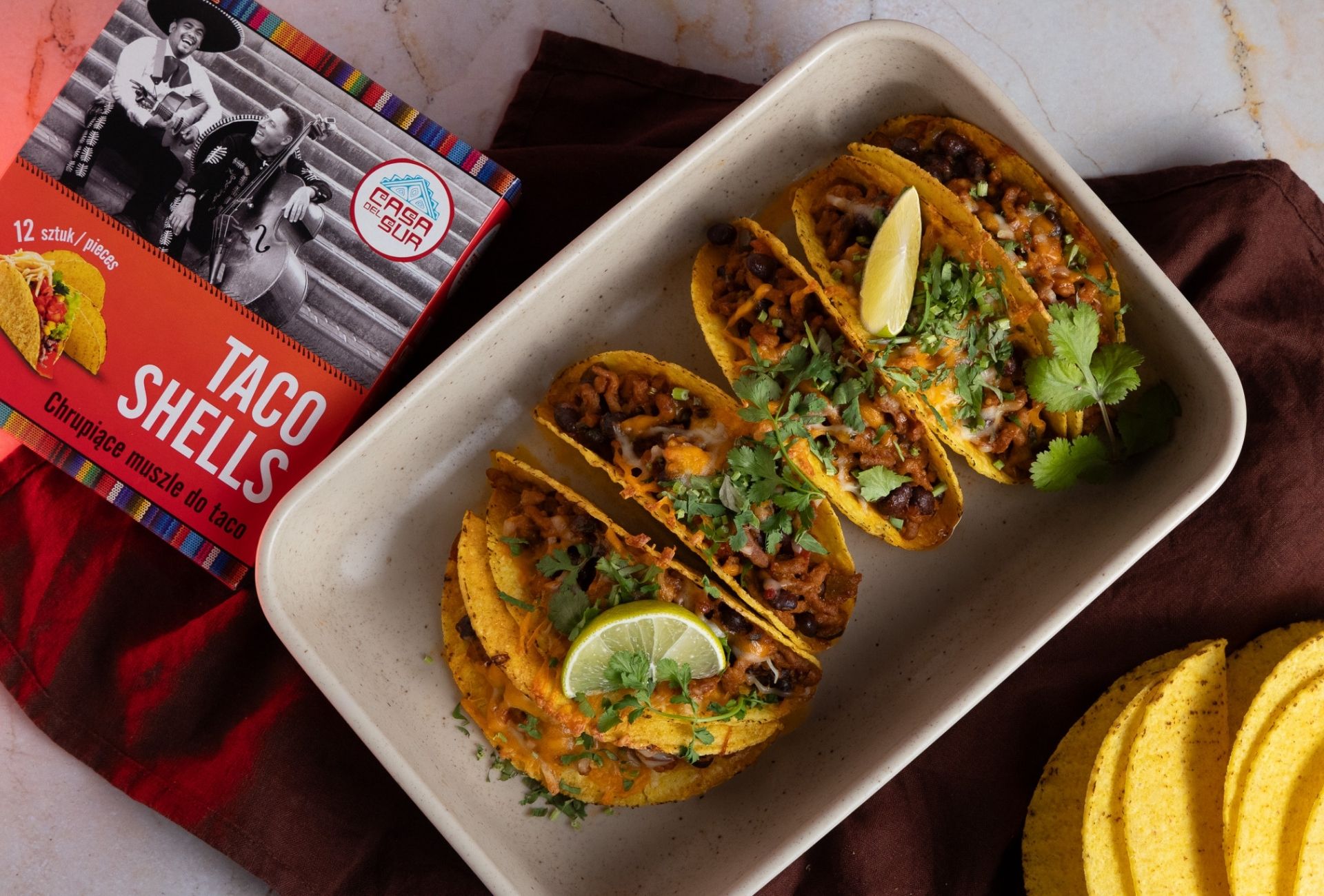 Danie z tacos wypełnionymi mięsem i serem w ceramicznej brytfannie, obok opakowania muszli taco marki Casa del Sur i stosu pustych tacos.